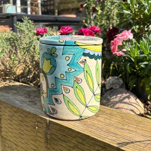 May include: A ceramic jar with a lid, featuring a colourful abstract design in shades of blue, green, yellow, and white. The jar has a whimsical, hand-painted style.