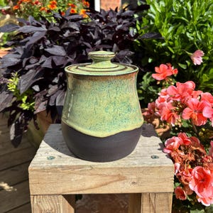 May include: A ceramic jar with a lid featuring a two-tone glaze. The upper portion and lid are finished in a mottled light green glaze, while the lower portion is a solid matte black. The jar has a rounded body and a small knob on the lid.