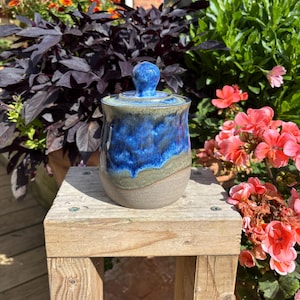 May include: A ceramic jar with a lid featuring a spherical knob. The upper portion of the jar is glazed in a vibrant, deep blue with a dripped effect, while the lower section transitions into a muted, earthy grey-brown tone with a smooth finish.