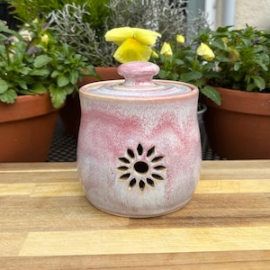 May include: A ceramic jar with a lid, featuring a floral cut-out design. The jar has a pink and white marbled glaze. The jar is approximately 10 cm tall and 10 cm wide.