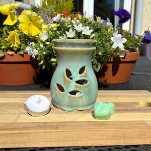May include: A teal ceramic essential oil diffuser with leaf-shaped cutouts. A white tea light candle and a green wax melt are next to the diffuser. The background includes potted flowers.