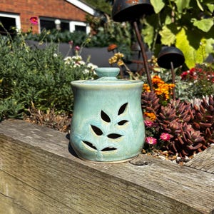 May include: A light green ceramic jar with a matching lid, featuring a cutout design of a leaf pattern on the front. The jar has a smooth, glossy glaze with subtle variations in colour and a slightly flared rim at the top of the vessel.