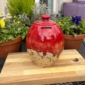 May include: A red ceramic money box with a glossy finish, featuring a slot for coins. The lower half of the bank has a speckled, tan colour. The money box is placed on a wooden chopping board, with potted plants in the background.