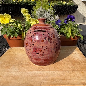 May include: A round, ceramic money bank with a lid, in shades of red and brown with a speckled pattern. The bank has a slot for inserting money. It sits on a wooden surface, with potted plants and flowers in the background.