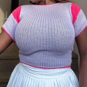 May include: A lilac-colored, short-sleeved knit top with bright pink accents on the shoulders and trim. The top is paired with a white pleated skirt, creating a casual, yet stylish ensemble. The knit fabric has a textured appearance.
