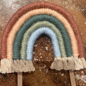 May include: A rainbow-colored yarn wall hanging with a light brown, peach, green, light green, light blue, and dark blue color scheme. The rainbow is attached to two wooden dowels with tassels at the bottom.