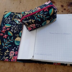 May include: A notebook and pencil case set with a floral pattern in red, pink, teal, and white on a navy blue background. The pencil case has a pink zipper. The notebook is open, revealing lined paper with handwritten text.