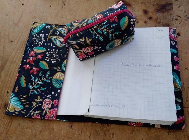 May include: A notebook and pencil case set with a floral pattern in red, pink, teal, and white on a navy blue background. The pencil case has a pink zipper. The notebook is open, revealing lined paper with handwritten text.