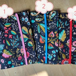 May include: Three fabric-covered journals with floral patterns and elastic closures. The journals feature a dark navy background with colorful flowers, leaves, and berries. Each journal has a different colored elastic band: pink, blue, and red.