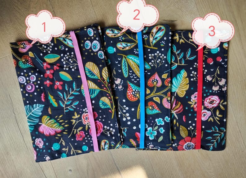 May include: Three fabric-covered journals with floral patterns and elastic closures. The journals feature a dark navy background with colorful flowers, leaves, and berries. Each journal has a different colored elastic band: pink, blue, and red.
