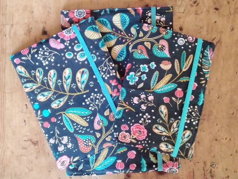 May include: Three notebooks with floral patterns in pink, teal, and gold on a dark blue background. Each notebook has a teal elastic band closure. The notebooks are arranged on a wooden surface.