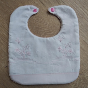 Może przedstawiać: Biały śliniaczek dla dziecka z różowym haftowanym kwiatowym wzorem i dwoma różowymi zatrzaskami. Śliniaczek ma zakrzywiony dekolt i prostokątny korpus z różową dolną krawędzią. Haft zawiera kwiaty i motyle.