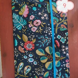 May include: A floral patterned journal cover with a dark blue background. The cover features a vibrant design of pink, teal, and gold leaves and flowers. A bright blue elastic band secures the journal. The number 9 is in a white cloud-shaped tag.