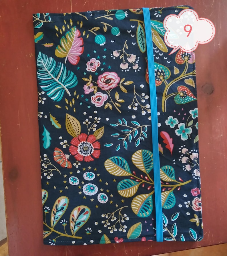 May include: A floral patterned journal cover with a dark blue background. The cover features a vibrant design of pink, teal, and gold leaves and flowers. A bright blue elastic band secures the journal. The number 9 is in a white cloud-shaped tag.