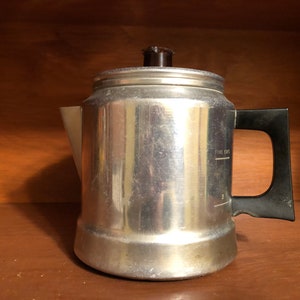 May include: A vintage aluminum coffee percolator with a black handle and a brown lid. The side of the pot has markings for five cups. The percolator is sitting on a wooden surface.