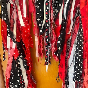Puede incluir: Una guirnalda decorativa hecha de tiras de tela colgantes en rojo, negro y blanco. Las tiras presentan varios patrones, incluyendo lunares blancos sobre tela negra, cintas s&oacute;lidas rojas y blancas, y tela roja con peque&ntilde;as siluetas de rat&oacute;n negras.