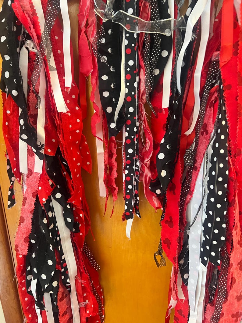 Puede incluir: Una guirnalda decorativa hecha de tiras de tela colgantes en rojo, negro y blanco. Las tiras presentan varios patrones, incluyendo lunares blancos sobre tela negra, cintas s&oacute;lidas rojas y blancas, y tela roja con peque&ntilde;as siluetas de rat&oacute;n negras.