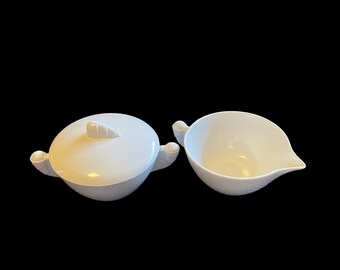 May include: A white ceramic sugar bowl and creamer set with a lid and handles. The sugar bowl has a small knob on the lid.