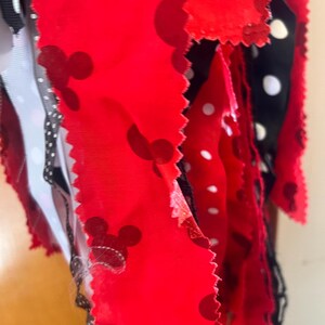 Puede incluir: Una colecci&oacute;n de tiras de tela con un fondo rojo y siluetas de cabezas de Mickey Mouse en rojo oscuro, intercaladas con tiras de tela negra decoradas con lunares blancos. Los bordes de la tela tienen un acabado dentado y recortado.
