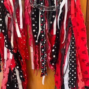 Puede incluir: Una guirnalda decorativa de tela compuesta por tiras colgantes en rojo, negro y blanco. Las tiras incluyen colores s&oacute;lidos, lunares blancos sobre tela negra y un patr&oacute;n de tela roja con peque&ntilde;as siluetas repetidas de cabezas de rat&oacute;n.