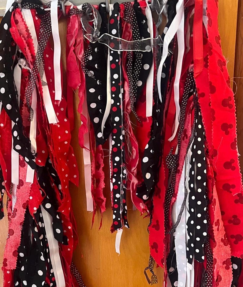 Puede incluir: Una guirnalda decorativa de tela compuesta por tiras colgantes en rojo, negro y blanco. Las tiras incluyen colores s&oacute;lidos, lunares blancos sobre tela negra y un patr&oacute;n de tela roja con peque&ntilde;as siluetas repetidas de cabezas de rat&oacute;n.