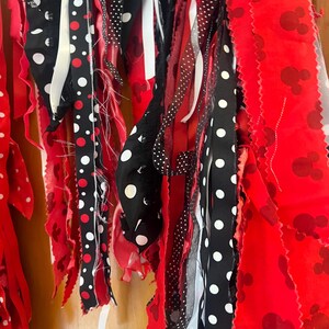 Puede incluir: Una guirnalda decorativa de tela con tiras colgantes en rojo, negro y blanco. Las tiras incluyen tela roja lisa, tela roja con un patr&oacute;n repetitivo de siluetas de cabeza de rat&oacute;n y cintas negras con lunares blancos de diversos tama&ntilde;os.