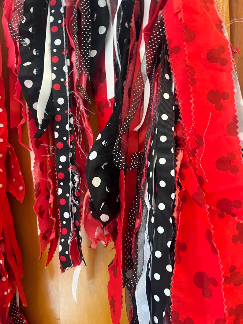 Puede incluir: Una guirnalda decorativa de tela con tiras colgantes en rojo, negro y blanco. Las tiras incluyen tela roja lisa, tela roja con un patr&oacute;n repetitivo de siluetas de cabeza de rat&oacute;n y cintas negras con lunares blancos de diversos tama&ntilde;os.