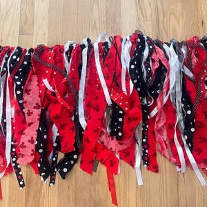 Puede incluir: Una guirnalda de tela con tiras rojas, negras y blancas. La tela incluye colores s&oacute;lidos en rojo, negro y blanco, junto con patrones de lunares blancos sobre tela negra y peque&ntilde;as siluetas de rat&oacute;n rojas sobre tela de color rojo.