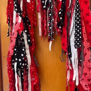 Puede incluir: Una guirnalda de tela con largas tiras de tela colgantes en rojo, negro y blanco. Los patrones incluyen lunares blancos sobre negro, lunares negros sobre blanco y tela roja con un peque&ntilde;o motivo repetitivo de siluetas de rat&oacute;n negras.