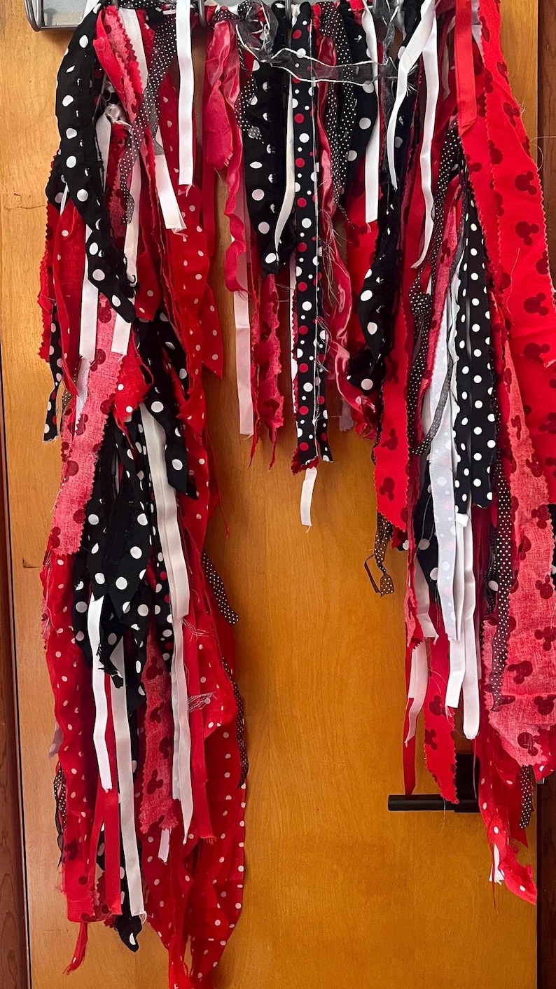 Puede incluir: Una guirnalda de tela con largas tiras de tela colgantes en rojo, negro y blanco. Los patrones incluyen lunares blancos sobre negro, lunares negros sobre blanco y tela roja con un peque&ntilde;o motivo repetitivo de siluetas de rat&oacute;n negras.