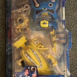 May include: A yellow and blue toy action figure called Max Steel Combat Diver Battle Gear. The toy comes with a water blasting weapon, a blue helmet, and a yellow submarine. The packaging includes the text "Water Blasting Weapon!" and a warning that the toy is not for children under 3 years old.