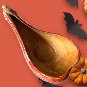 May include: A ceramic serving dish shaped like a gourd, with a glossy brown interior and orange-brown exterior. The dish is decorated with a pumpkin and black bat cutouts on an orange background, perfect for Halloween.