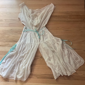 May include: A vintage white romper with a sleeveless design and lace trim. The garment features a ruffled bodice and wide legs, tied with a light blue ribbon. The fabric appears lightweight and flowing, suggesting a delicate, airy feel.