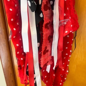 Puede incluir: Una guirnalda decorativa de tela con tiras colgantes en rojo, negro y blanco. El surtido incluye cintas rojas lisas, cintas negras con lunares blancos, tela roja con siluetas de cabezas de rat&oacute;n y cintas de color blanco.