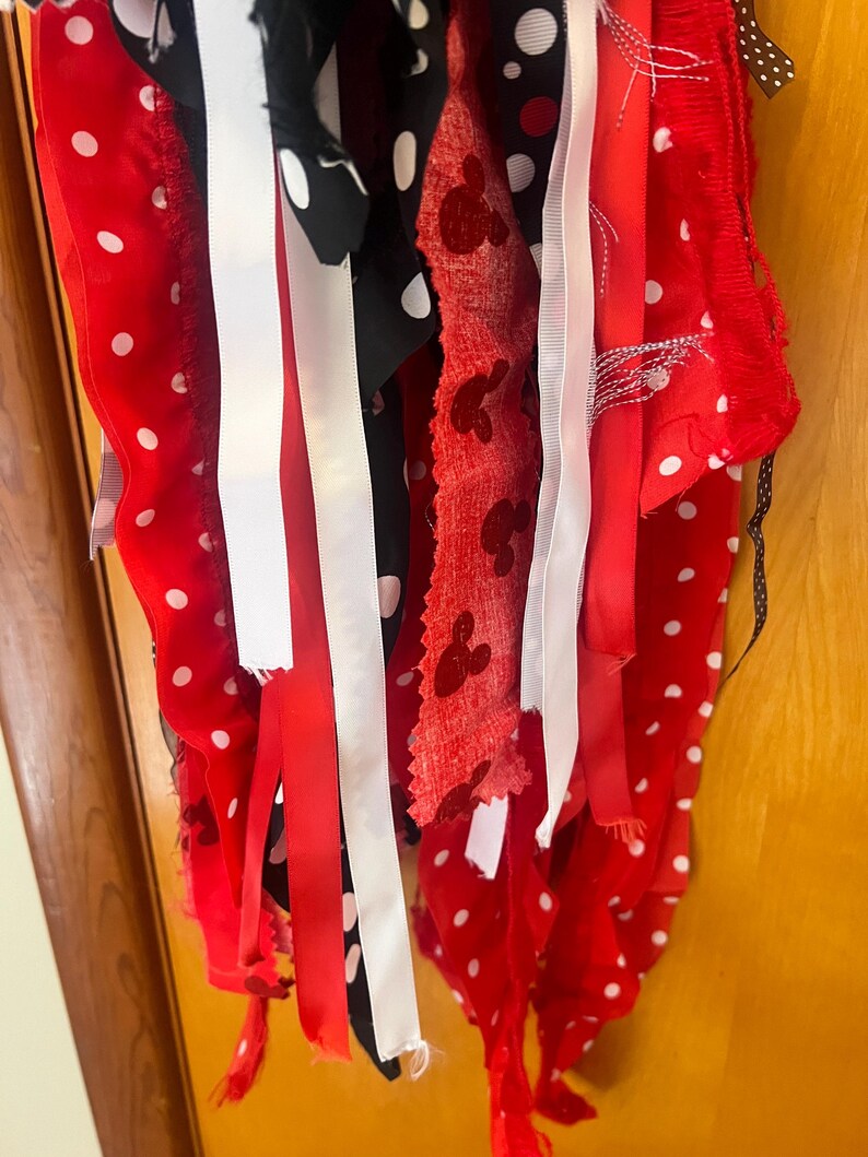 Puede incluir: Una guirnalda decorativa de tela con tiras colgantes en rojo, negro y blanco. El surtido incluye cintas rojas lisas, cintas negras con lunares blancos, tela roja con siluetas de cabezas de rat&oacute;n y cintas de color blanco.