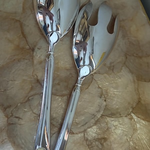 May include: A pair of polished silver-tone salad serving utensils consisting of one solid spoon and one three-pronged fork. Each handle features a smooth, rounded design with a small circular sticker near the base that reads Made in China.