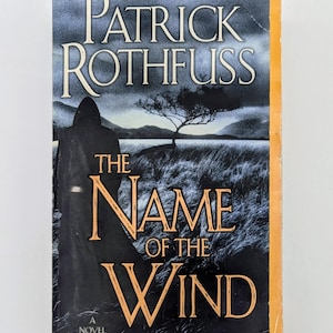 May include: A paperback book titled The Name of the Wind by Patrick Rothfuss. The cover features a dark, hooded figure standing in a grassy field at dusk, with a lone tree against a cloudy sky. The title is printed in large, gold-colored lettering.