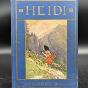 May include: A vintage hardcover book titled "HEIDI" from the Windermere Series. The cover is blue with gold lettering and a framed illustration of a girl running on a path. The girl wears an orange dress and carries a basket.