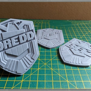 May include: Three gray 3D printed badges with a stylized eagle design. The first badge says "DREDD", the second says "NAME HERE", and the third says "BAIN".