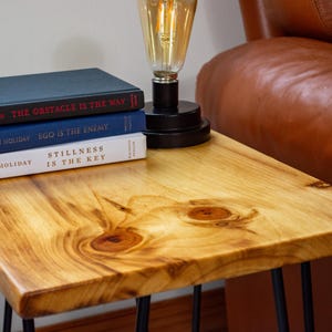 Pode incluir: Uma mesa lateral de madeira com acabamento natural, com uma pilha de livros e uma lâmpada. Os livros são intitulados "THE OBSTACLE IS THE WAY", "EGO IS THE ENEMY" e "STILLNESS IS THE KEY". A lâmpada tem uma base preta e uma lâmpada de estilo vintage.