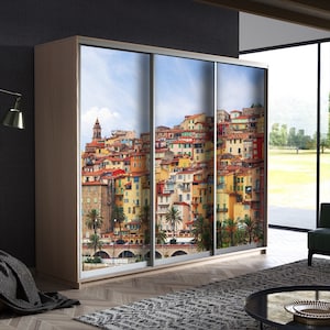 May include: A three-door wardrobe with a light wood frame. The sliding doors feature a vibrant image of a coastal town with colorful buildings, palm trees, and a blue sky. The image is a scenic view of a European town.