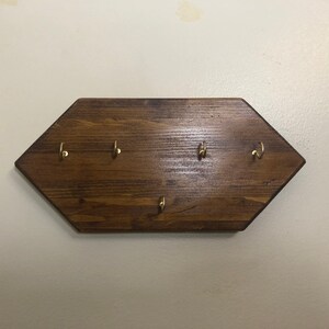 Puede incluir: Un porta llaves de madera hexagonal con cinco ganchos de latón. El porta llaves está teñido de un color marrón oscuro.