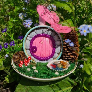 May include: A miniature fairy door with a pink door and purple trim, surrounded by small mushrooms, a seashell, pine cones, and a pink butterfly on a white saucer.