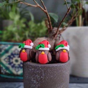 May include: Three small, brown bird figurines with red breasts and Santa hats, perched on a gray pot. The hats are red with white trim and green holly. The birds have yellow beaks and are set against a blurred background of green plants.