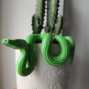 Könnte beinhalten: Eine grüne Keramikschlange, die sich um einen weißen Blumentopf mit Schneeflockenmustern windet. Die Schlange hat weiß-grüne Streifen. Ein grüner Kaktus befindet sich im Topf.