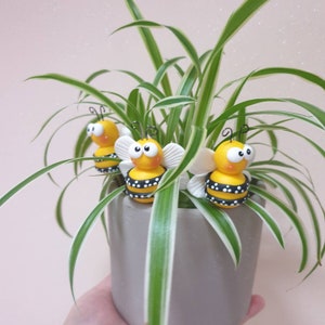 Könnte beinhalten: Drei gelbe und schwarze Hummeln mit weißen Flügeln sind in einer grünen Topfpflanze eingebettet. Die Bienen haben schwarze Augen und sind aus Ton gefertigt.