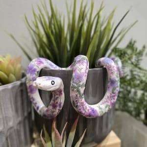 May include: A decorative snake figurine with a floral pattern in shades of purple and white. The snake is coiled around a grey pot containing green plants. The figurine has a white head with a black eye.
