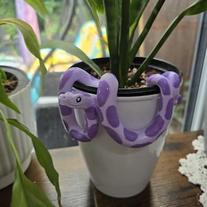 May include: A flexible, light purple snake figurine with darker purple oval spots and black-and-white eyes, coiled around the rim of a plain white cylindrical plant pot containing a green plant with long, pointed leaves.