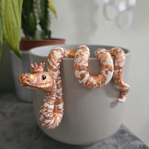 Könnte beinhalten: Dekorative Schlangenfigur mit kupfer-weißem Muster, die eine kleine kupferfarbene Krone trägt. Die Schlange ist über den Rand eines grauen Keramiktopfes drapiert. Der Hintergrund umfasst grüne Pflanzen und eine weiße Wand.