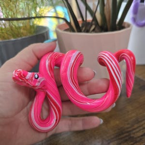 May include: A coiled snake figurine featuring a marbled pattern of vibrant hot pink, white, and red swirls. The sculpture has a smooth, glossy finish with a defined head, a pointed tail, and small black and white painted eyes on the sides of its head.